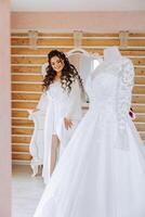 A happy bride is preparing for her luxurious wedding in a hotel room, with a wedding dress on a mannequin nearby. Portrait of a woman with fashionable hair, makeup and a smile in a dressing gown. photo