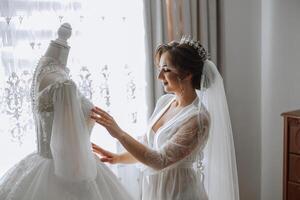 A happy bride is preparing for her luxurious wedding in a hotel room, with a wedding dress on a mannequin nearby. Portrait of a woman with fashionable hair, makeup and a smile in a dressing gown. photo