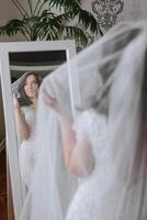 un hermosa novia en un elegante Boda vestir soportes en frente de un espejo debajo un velo en su habitación en el Mañana. foto