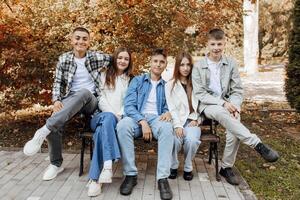 un grupo de adolescentes tener divertido y alegrarse mientras sentado en un parque banco. Adolescente compañeros de clase son descansando en contra el antecedentes de otoño naturaleza. foto