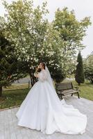 Curly brunette bride in a lace dress with an open bust, in a lush veil holds a bouquet and poses against the background of green trees. Spring wedding photo