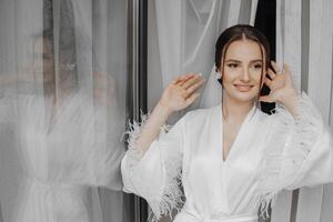 portrait of bride girl in white gown with professional hairstyle and natural makeup in hotel room with reflection in window. The best day photo