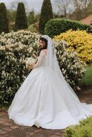 Rizado morena novia en un lozano velo y manga larga vestir poses siguiente a un blanco lila. hermosa pelo y constituir. primavera Boda foto