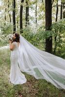 A beautiful bride is standing in a green spring forest in a glamorous white wedding dress with a view of the veil. A romantic bride in a white dress is posing against the background of the forest. photo