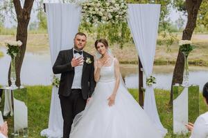 novias y novios lucir su Boda anillos cerca un blanco arco decorado con flores durante un Boda ceremonia. el novia es participación un ramo. primavera boda. contento momentos foto