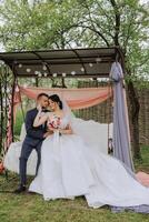 A beautiful young bride in a spring park embraces her groom while sitting on a swing in the garden. Beautiful wedding white dress with a long train. Walks in the park. A happy and loving couple. photo