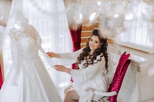 A happy bride is preparing for her luxurious wedding in a hotel room, with a wedding dress on a mannequin nearby. Portrait of a woman with fashionable hair, makeup and a smile in a dressing gown. photo