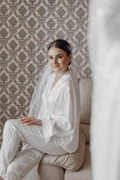The bride in a silk suit and veil poses in her room, sitting on the couch. Morning of the bride. Preparation. Wedding photography photo