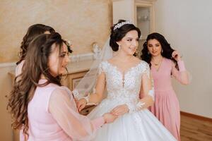 A friend helps the bride fasten her dress. A woman helps her friend fasten the buttons on the back of her wedding dress. The morning of the bride, the creation of a family, an important event. photo