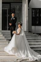 The bride and groom are walking near the hotel and posing, happy and enjoying the day, holding hands. A long train on the dress. Winter wedding photo