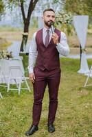 un joven, barbado hombre en un borgoña traje habla dentro un micrófono durante un Boda ceremonia. presentador a el boda. negocio estilo. foto