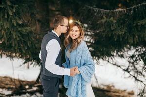 contento y sonriente novia y novio en el antecedentes de invierno bosque. un hermoso novio tiernamente sostiene el manos de su novia y Besos su. el concepto de un moderno invierno boda. foto