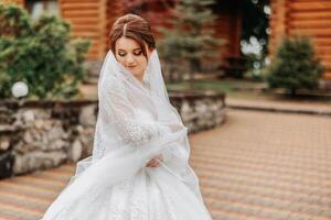 A red-haired bride twirls her long veil against a background of wooden houses. Magnificent dress with long sleeves, open bust. Summer wedding photo