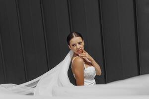 Beautiful young bride girl wearing a white dress with a long veil and posing near a gray wall in a modern city. photo