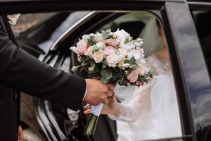 un hermosa novia, sentado en un auto, da su mano a su marido. un hermosa novia con un ramo de flores de flores en su manos es sentado en un elegante costoso coche. foto