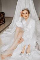 A bride in a silk suit with a veil poses in her room, sitting on the floor next to her wedding dress. Morning of the bride. Preparation photo