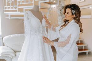 Morning of the bride before the wedding. A beautiful young woman with a veil and a beautiful hairstyle in a white robe near her wedding dress on a mannequin. Professional makeup. Last preparations. photo