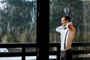 Side view of a man in suspenders wearing a bow tie while standing on a balcony overlooking snow-capped mountains. Rest in the mountains. Mountain air. Wedding in winter photo