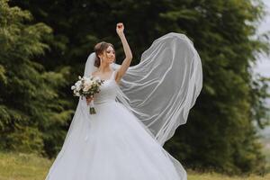 The bride in a lush dress and veil, holding a bouquet of white flowers and greenery, poses with the veil thrown in the air, against the background of green trees. Spring wedding photo