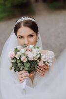 brunette bride in a lacy white dress with a long train, posing with a bouquet of white and pink flowers, against the background of trees. The veil is in the air. Beautiful hair and makeup. photo