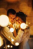 a happy bride and groom embrace in a dark room illuminated by defocused lights of a garland photo