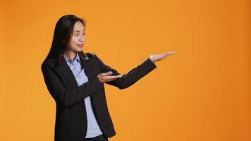 Asian employee points at left or right sides in studio, creating new web commercial over orange background. Young business manager working on marketing advertisement during shoot. video