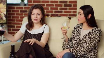 asiatico donna chat con amico su confortevole divano a appartamento festa, potabile vino e godendo conversazione. gioioso bff attraente su con ogni altro a casa, consumando Champagne video