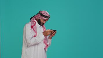 Islamic guy playing fun mobile videogames on phone, dressed in traditional gown with headscarf. Middle eastern person engaging in online gaming contest using smartphone app. video