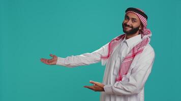 Confident islamic guy in national costume showcasing with hands at copyspace for advertisement commercial on camera. Joyful arab person presenting sides with arms in studio. video