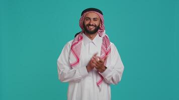 Muslim adult in traditional gown showing support while applauding with hands in studio. Middle eastern guy wearing white thobe and checkered headscarf, clapping and cheering. video