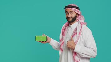 Muslim model with greenscreen layout on mobile phone posing with confidence in studio, showcasing isolated mockup chromakey on smartphone screen. Young adult presents blank copyspace display. video