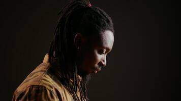 African american guy looking at camera with confidence, feeling happy and relaxed with dreads and cool clothing items. Model posing, standing over black background smiling. Handheld shot. video