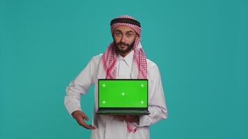 Islamic man presents greenscreen on pc, showing isolated chromakey template on laptop display. Young person holding wireless computer with mockup screen and copyspace, wearing thobe and headscarf. video