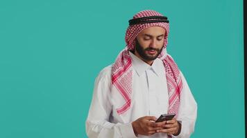 sorridente adulto digitando mensagens em telefone, vestindo muçulmano tradicional roupas. islâmico árabe pessoa vestido dentro branco Thobe e xadrez ghutra conversando em social meios de comunicação página da Internet, mensagens de texto. video