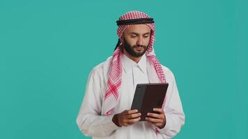 Middle eastern guy browsing tablet on camera, navigating webpages online on digital device. Male model in arabic clothing using gadget to check internet sites, texting on social media. video