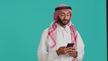 musulman homme dans traditionnel tenue envoyer des SMS sur mobile téléphone tandis que posant fièrement dans studio. milieu est adulte habillé dans thobe et à carreaux foulard, Envoi en cours en ligne messages sur téléphone. video