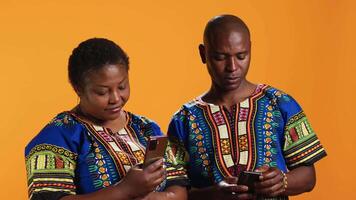 étnico casal rolagem em social meios de comunicação contas dentro estúdio, usando smartphones para navegar conectados rede paginas web. africano americano pessoas dentro tradicional vestuário verificação Internet sites. video