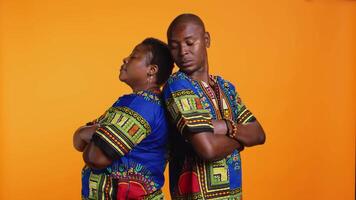 Serious proud ethnic couple standing back to back in studio, posing with confidence over orange background. African american man and woman dressed in traditional clothing, carefree partners. video