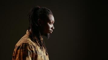 Smiling person posing in studio with black background, feeling confident wearing dreads and camo clothing. Cool guy with trendy hair acting stylish on camera, attractive man. Handheld shot. video