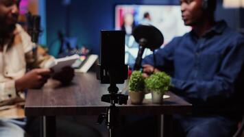 Focus on smartphone on tripod used by podcast host in blurry background to record guest telling interesting story to fans. Woman invited in studio sharing facts, filmed by phone, close up panning shot video