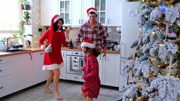 contento tradicional familia de papá, mamá, hija en festivo Papa Noel sombreros gracioso y divertido bailando en un blanco cocina con un Navidad árbol y decoración. nuevo año, familia valores. video