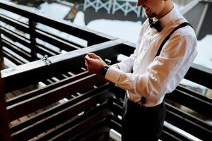 a young man wearing suspenders, a white shirt and a black bow tie puts on his watch. A wedding in the mountains in winter photo