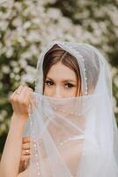 Pelirrojo novia en un lozano vestir con un abierto busto, posando envuelto en un velo, en contra el antecedentes de floración arboles primavera Boda en naturaleza. foto