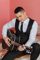 The groom sings songs to the guitar in the morning in his room. Groom in a black suit. Happy man. Preparation for the wedding. photo