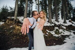 A young couple, the bride and groom, smilingly show off their wedding rings against the background of tall trees. Have a good time laughing. Winter wedding photo