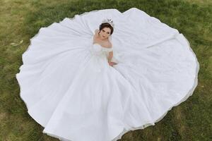 brunette bride in off-the-shoulder lace white dress and tiara posing with a bouquet of white and pink flowers while sitting on green grass. Beautiful hair and makeup. Spring wedding photo