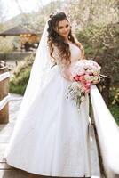 Curly brunette bride poses with a bouquet, near a wooden railing. Magnificent dress with long sleeves, open bust. Spring wedding photo