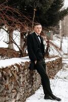 Wedding portrait of the groom. The groom is standing near a stone fence. A man in a black suit. Winter wedding photo