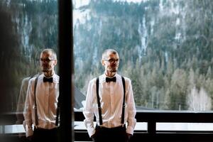 Window view of a man in suspenders with a bow tie standing on a balcony overlooking snow-capped mountains. Rest in the mountains. Mountain air. Wedding in winter photo