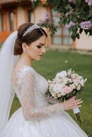 brunette bride in a lacy white dress with a long train, posing with a bouquet of white and pink flowers, against the background of trees. The veil is in the air. Beautiful hair and makeup. photo